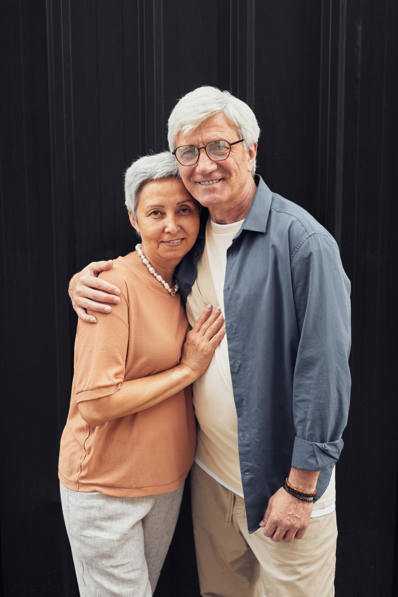 Vertical portrait of modern mature couple embracing and looking at6 camera while standing against black background