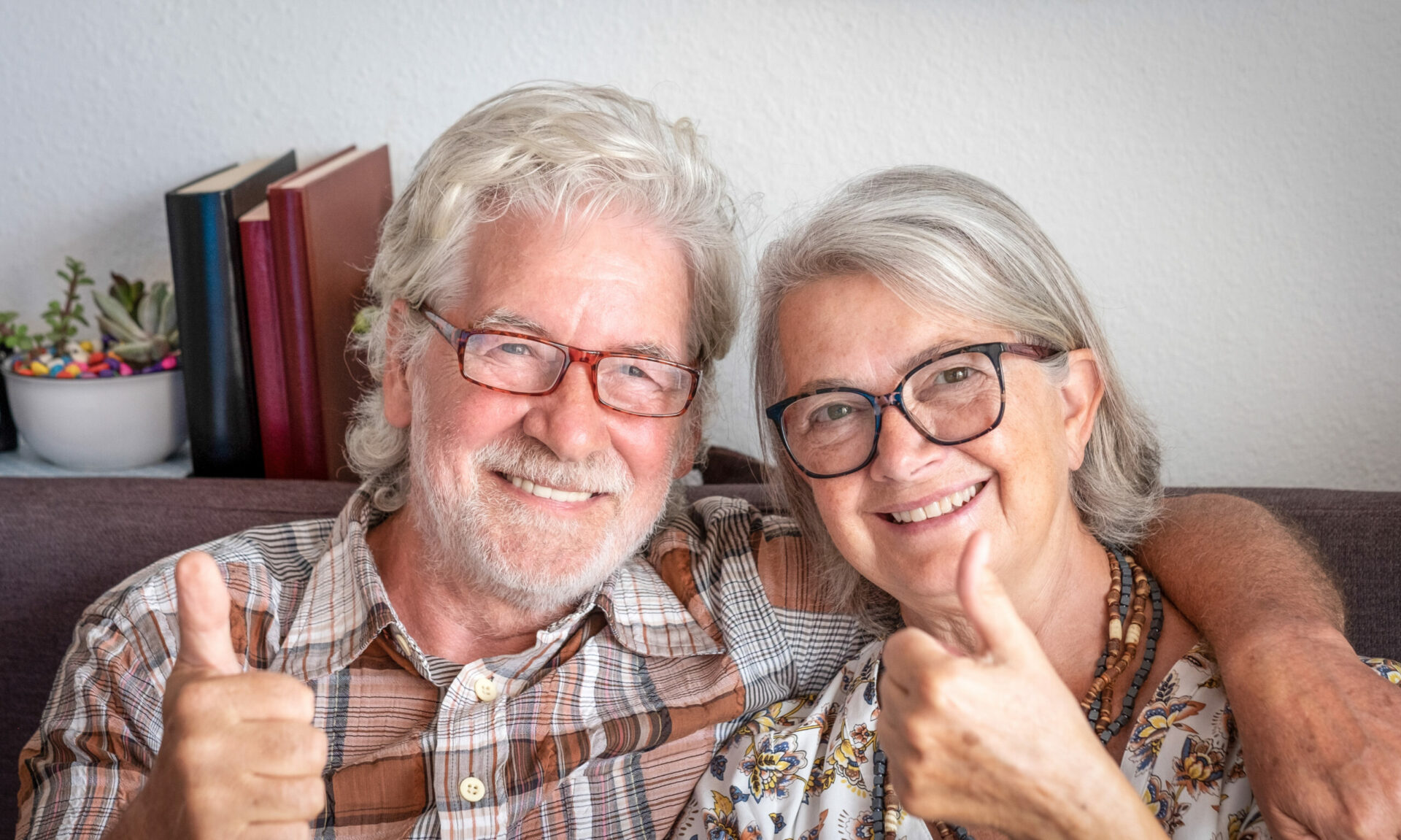 Portrait of caucasian smiling adult senior couple sitting at home on sofa looking at camera doing positive gesture with thumb up. Elderly white haired people enjoying together good time and retirement.